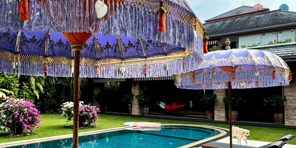 Handcrafted Balinese parasols in lavender with golden details and fringe, elegantly placed by a luxurious poolside garden. The scene includes vibrant flowers, a clear swimming pool, and a relaxing outdoor ambiance.